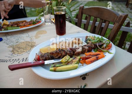 Ein griechisches Souvlaki mit Gemüse und frischer Cola in einem hohen Glas. Die verheiratete Frau isst auch, sitzt auf Holzstühlen. Das Tischtuch ist fleckig. Stockfoto