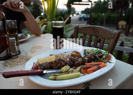 Ein griechisches Souvlaki mit Gemüse und frischer Cola in einem hohen Glas. Die verheiratete Frau isst auch, sitzt auf Holzstühlen. Das Tischtuch ist fleckig. Stockfoto