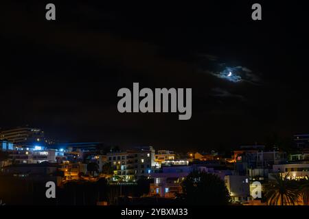 Mondsichel, der nachts in Teneriffa durch Wolken über einer beleuchteten Stadtlandschaft von Puerto de la Cruz glüht. Stockfoto