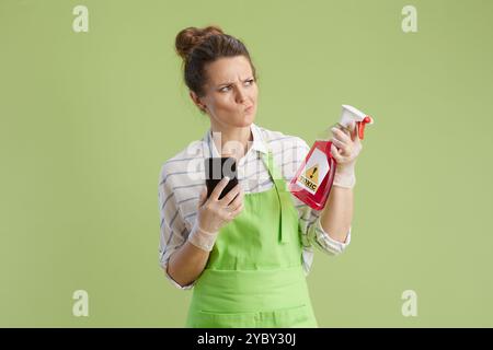 Frühjahrsputz. Besorgte moderne Frau in grüner Schürze und Gummihandschuhen isoliert auf grünem Hintergrund mit Smartphone und Flasche giftiges Reinigungsprodukt Stockfoto