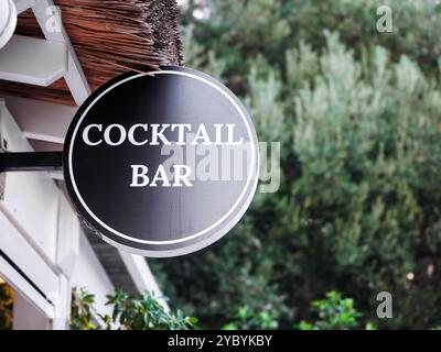 Rundes schwarz-weißes Schild für die Cocktailbar, das vor einem Gebäude mit Strohdach hängt Stockfoto