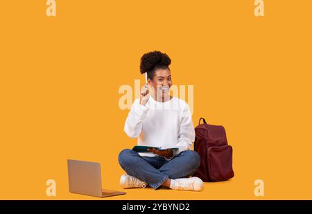 Glückliche junge Frau, die mit einem Laptop und Tablet vor orangefarbenem Hintergrund lernt Stockfoto