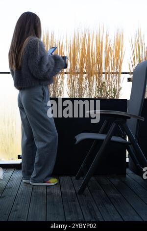 Eine junge Frau auf einer Terrasse im Herbst Prag, die Kaffee und Smartphone in einem grauen Kunstpelzmantel hält. Stockfoto