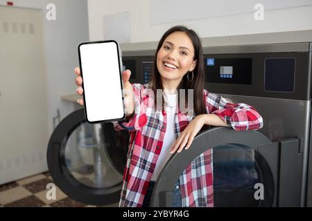Lächelnde Frau mit Smartphone in einem Waschsalon, während sie Wäsche macht Stockfoto
