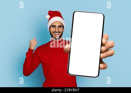 Fröhlicher Mann in einem Weihnachtsmütze zeigt Smartphone auf blauem Hintergrund Stockfoto