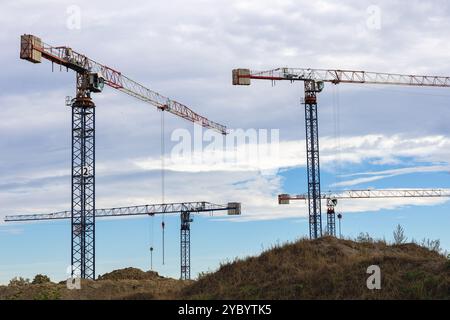 Turmkräne auf einer Baustelle gegen bewölkten Himmel und grasbewachsenes Gelände an einem sonnigen Tag Stockfoto