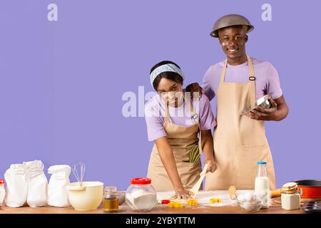Glücklicher Kerl und erschöpfte Dame, die von Kopf bis Fuß mit Mehl bedeckt sind, während sie auf violettem Hintergrund kochen Stockfoto