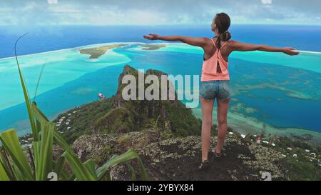 Die junge Frau steht mit gestreckten Armen auf einem Berggipfel und genießt den atemberaubenden Blick auf eine tropische Insellagune im südpazifik Stockfoto