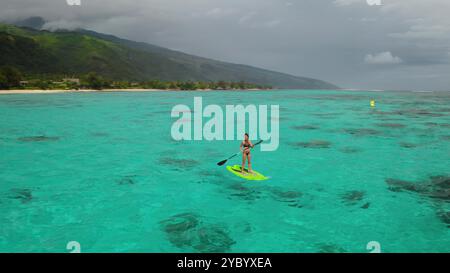 Aus der Vogelperspektive einer Frau, die Stand-Up-Paddle-Boarding in einer türkisfarbenen Lagune mit einer bergigen tropischen Insel im Hintergrund genießt Stockfoto