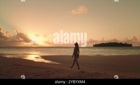 Frau genießt einen Spaziergang bei Sonnenuntergang an einem tropischen Strand auf den malediven. Die Silhouette der Frau ist vor der untergehenden Sonne zu sehen und schafft eine ruhige und friedliche Atmosphäre Stockfoto