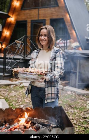 Frau, die Spieße für das Grillen am offenen Feuer vorbereitet, genießt Outdoor-Kochen mit einem Lächeln Stockfoto