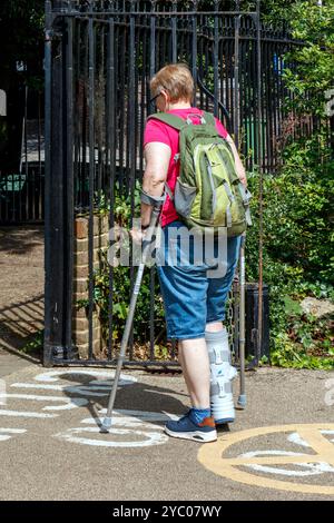 Eine Frau mit gebrochenem Bein in einem Schutzstiefel auf Krücken in einem Park, London, Großbritannien Stockfoto