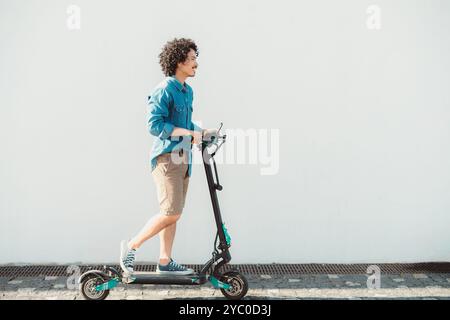 Junger asiatischer Mann, der auf einem Elektroroller fährt, ein Jeansshirt, khakifarbene Shorts und Turnschuhe trägt, vor einem hellen, minimalistischen Hintergrund. Seitenansicht auf einem Stockfoto