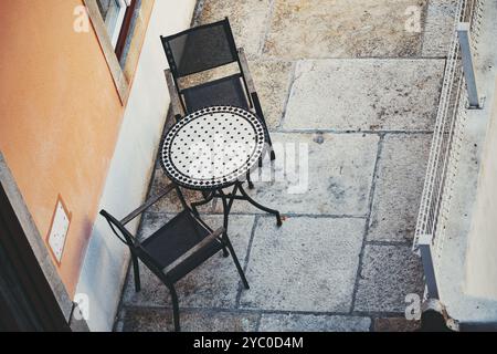 Tele-Blick von oben auf ein kleines Café im Freien mit einem runden Mosaiktisch und zwei schwarzen Mesh-Stühlen auf einer Steinterrasse. Die Szene fängt ein gemütliches ein, mi Stockfoto