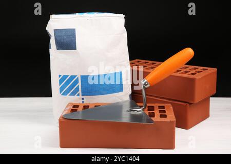 Zementpulver in Beutel, Kelle und roten Ziegeln auf weißem Holztisch. Baumaterialien und Bauwerkzeuge Stockfoto