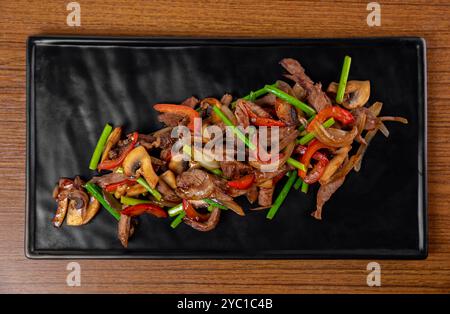 Gebratenes Schweinefleisch und Rindfleisch mit Knoblauchpfeilen, Pilzen und Gemüse auf einem schwarzen Teller auf einem Holztisch Stockfoto