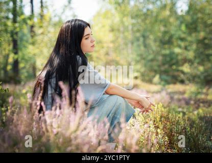 Eine junge Frau sitzt ruhig auf dem Waldboden, ihr langes Haar zieht sich über den Rücken. Sie erscheint tief in Gedanken, umgeben von blühenden Wildblumen Stockfoto