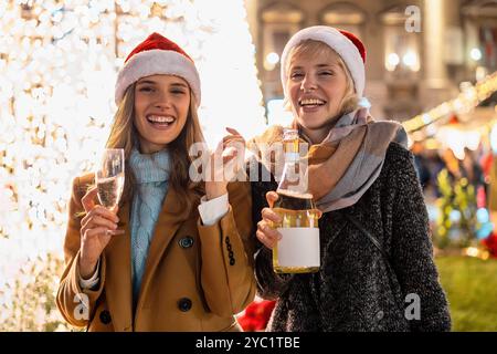 Zwei fröhliche Frauen in Weihnachtsmützen genießen Weihnachtsmarktfeste. Freunde teilen sich Champagner und Lachen unter funkelnden Lichtern. Warme Atmosphäre Captu Stockfoto