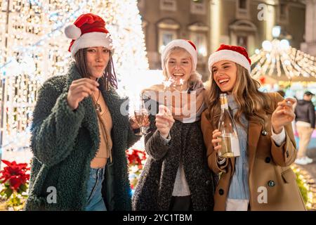 Drei verschiedene Frauen mit Weihnachtsmützen genießen Weihnachtsmarktfeste. Freunde verschiedener Ethnien und Alters halten Sparkler und Champagner und lachen Stockfoto