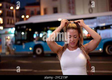 Die junge Frau bindet sich die Haare in einem Pferdeschwanz, während sie nachts auf einer Stadtstraße steht, mit einem Bus im Hintergrund Stockfoto