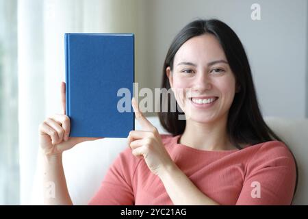 Glückliche asiatische Frau, die vor der Kamera einen schwarzen Buchdeckel zeigt, der zu Hause nachgeahmt wird Stockfoto