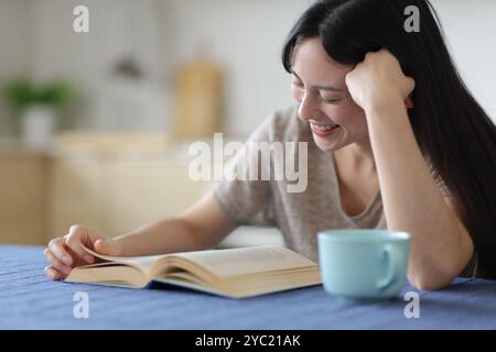 Glückliche asiatische Frau, die ein Papierbuch in der Küche zu Hause liest Stockfoto