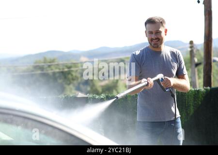 Glücklicher Mann, der Auto mit Hochdruckschlauch im Garten zu Hause wäscht Stockfoto