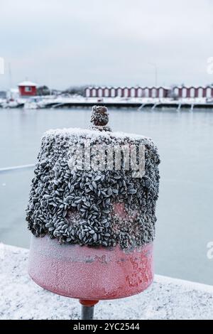 Am Wasserrand steht eine Boje, beschichtet mit einer Schicht aus Seebrücken und Schnee, mit einem gefrorenen Hafen und roten Bootshäusern, die während einer im Hintergrund sichtbar sind Stockfoto