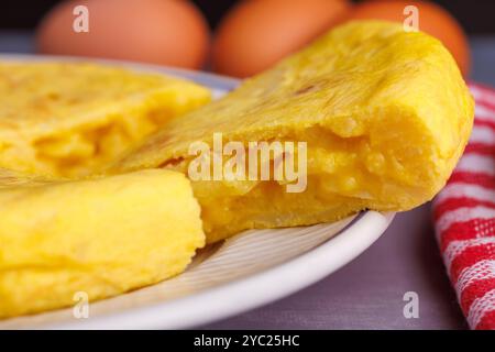 Ein detaillierter Blick auf ein spanisches Kartoffelomelett, mit einem Stück neben einer karierten Serviette, Brot und Eiern auf einem bläulichen Betontisch Stockfoto