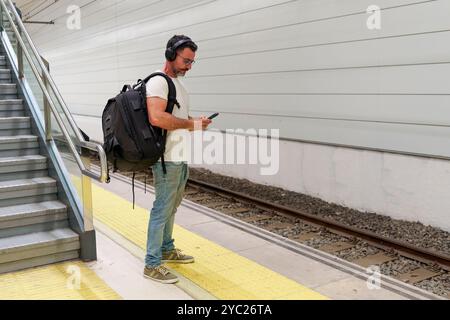 Mann mittleren Alters in lässiger Kleidung mit Rucksack mit Smartphone für E-Ticket während der Wartezeit auf den Zug am Bahnhof Stockfoto