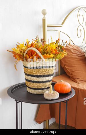 Wunderschön angeordneter Korb gefüllt mit gelben Herbstblättern und kleinem orangenem Kürbis auf schwarzem Tisch neben Bett mit Kissen. Herbstdekoration Stockfoto