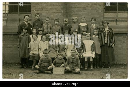 Original-Postkarte Schulhofporträt von Kleinkindern aus den 1920er Jahren. Schulkinder der 1920er Jahre. Eine Tafel davor trägt die Aufschrift „Colwich C.E. School Group 111“ und datiert vom 12. Juni 1924. Viele ernsthafte Charaktere, die verschiedene Moden tragen, viele Handwerker, stehen mit Lehrern. Colwich, in der Nähe von Stafford und Rugeley, Staffordshire, Großbritannien Stockfoto