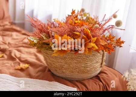 Schöne Anordnung von orange herbstfarbenen Blättern und Blumen wird in gewebtem Korb gezeigt. Der Korb liegt auf einem Bett in warmen Farbtönen und ergänzt das gemütliche Ambiente Stockfoto