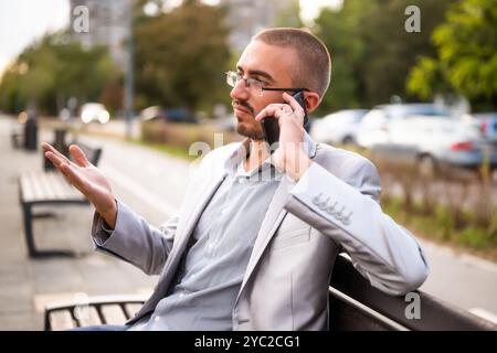 Porträt eines ungläubigen Geschäftsmannes, der am Telefon spricht. Mann, der in der Stadt auf der Bank sitzt und telefoniert. Stockfoto