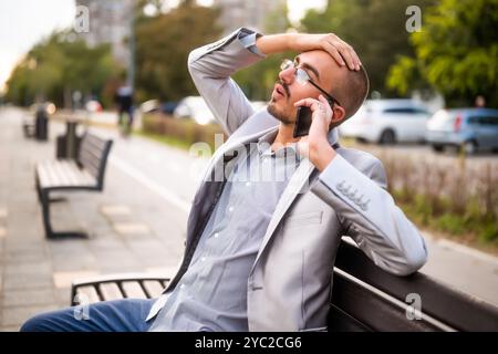 Porträt eines ungläubigen Geschäftsmannes, der am Telefon spricht. Mann, der in der Stadt auf der Bank sitzt und telefoniert. Stockfoto