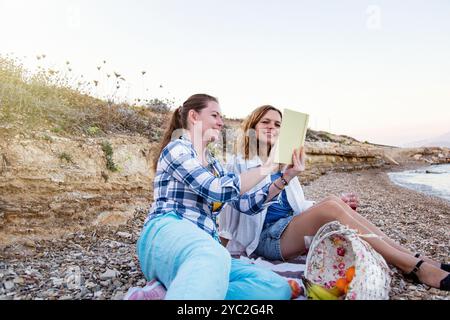 Zwei Frauen sitzen an einem felsigen Strand, schauen sich Bücher an und lächeln Stockfoto