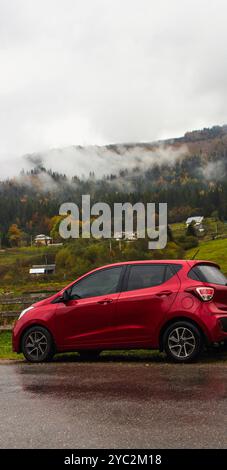 Rotes Auto auf der Straße in den Bergen an regnerischen Tagen. Autoverkehr im Herbst. Fließheckwagen in Bergdorf mit Wolken. Road Journey in Karpaten Stockfoto