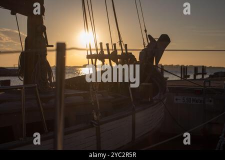 Ein historisches hölzernes Segelschiff legte bei Sonnenuntergang an und warf lange Schatten auf das Deck, während die Sonne unter dem Horizont untergeht. Die Silhouette der rigg des Schiffes Stockfoto