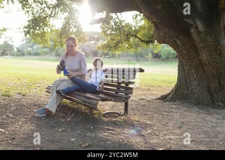 Mutter hilft ihrem Sohn, seine Schuhe anzuziehen, während sie auf einer Parkbank unter einem Baum sitzt Stockfoto