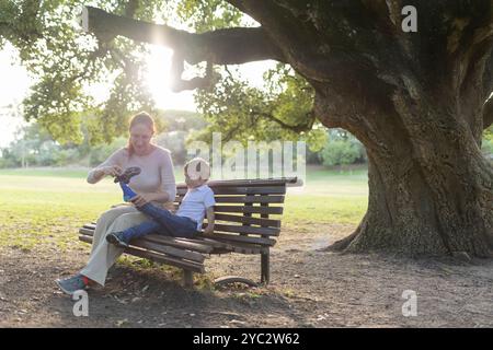 Mutter hilft ihrem Sohn, seine Schuhe anzuziehen, während sie auf einer Parkbank unter einem Baum sitzt Stockfoto