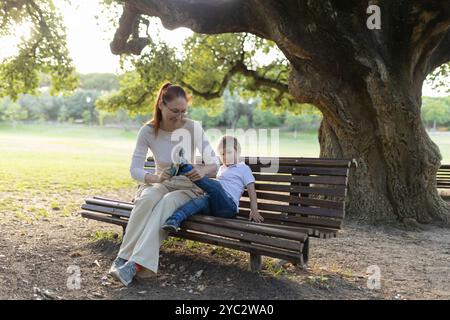 Mutter hilft ihrem Sohn, seine Schuhe anzuziehen, während sie auf einer Bank im Park sitzt Stockfoto