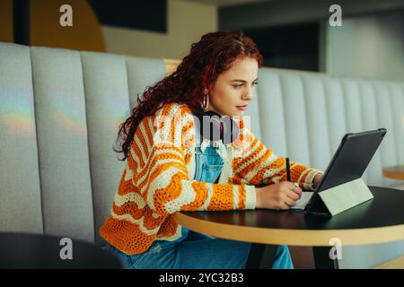 Eine Studentin studiert intensiv mit einem Tablet, trägt Kopfhörer und einen farbenfrohen gestreiften Pullover in einer gemütlichen, modernen Café-Umgebung. Stockfoto