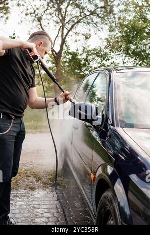 Mann, der ein Auto mit einem Hochdruckreiniger an einem sonnigen Tag in einer grünen Umgebung im Freien wäscht. Stockfoto