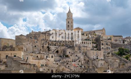 Eingebettet in die Hügel bietet Matera Italien einzigartige Höhlenhäuser und komplizierte Fassaden, die in sanftes Sonnenlicht getaucht sind. Der hoch aufragende Glockenturm ist Wächter über dieser historischen Stadt, während die Wolken über sich ziehen Stockfoto