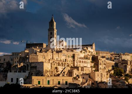 Eingebettet in die Hügel von Apulien, zeigt Matera seine alten Höhlenwohnungen, während goldenes Licht die historische Architektur durchflutt und einen beeindruckenden Kontrast zum stürmischen Himmel bei Sonnenuntergang schafft. Stockfoto
