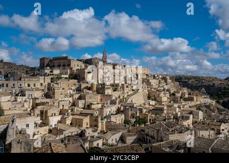 Eingebettet in sanfte Hügel, zeigt die historische Stadt Matera ihre einzigartige Steinarchitektur unter einem Hintergrund flauschiger Wolken. Diese bezaubernde aussicht zeigt jahrhundertealte Kultur und Kulturerbe. Stockfoto