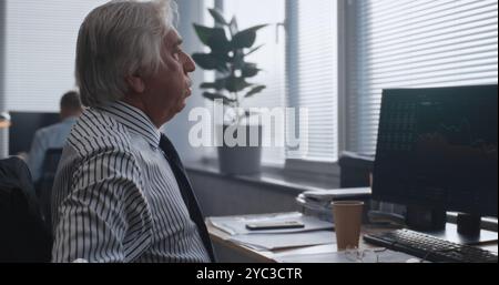 Müde Senior Financial Specialist analysiert Papiere und Dokumente, überwacht Börsen und Aktien am Computer. Der professionelle Händler arbeitet im modernen Büro der internationalen Börsengesellschaft. Stockfoto