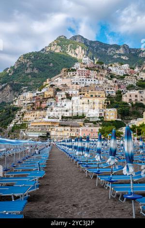 Farbenfrohe Strandstühle säumen die Sandstrände von Positano, mit malerischen Klippen, die hinter diesem Küstenjuwel ragen. Die lebendigen Häuser kaskadieren die Berge hinunter und bieten einen atemberaubenden Blick. Stockfoto