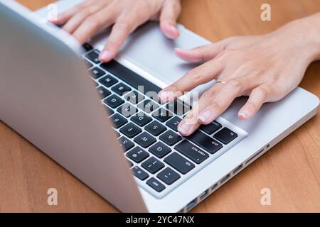 Nahansicht der Hände, die auf einer Laptop-Tastatur tippen. Der Laptop ist silberfarben und auf einem Holztisch. Die Finger befinden sich über den Tasten, IND Stockfoto