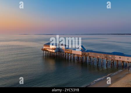 Sonnenlicht filtert durch die hölzernen Stützen eines Piers und wirft Reflexionen auf den nassen Sand darunter. Die ruhige Meereskulisse verstärkt den friedlichen geldautomaten Stockfoto